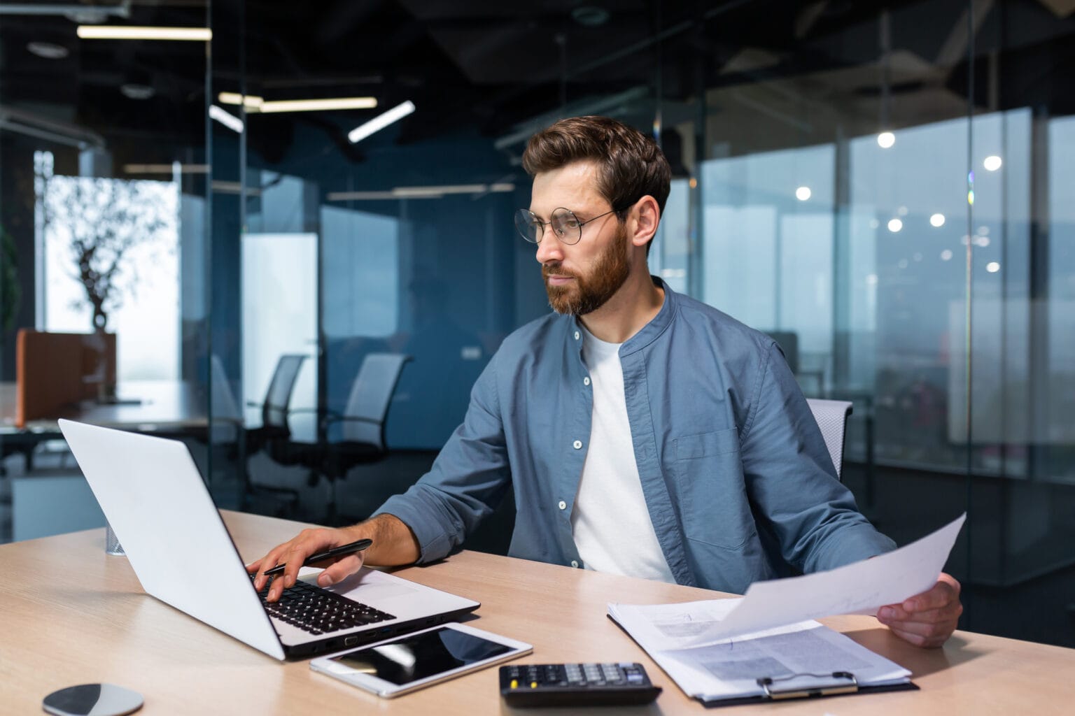 A man financier, auditor sits in the office . he holds documents, checks accounts. types on a laptop